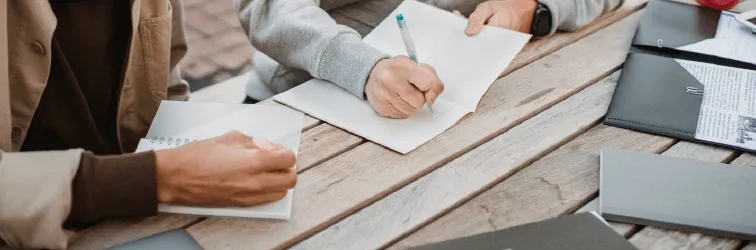 A photo of two students studying together at a wooden table outside. Only their hands are shown, and they are both writing on paper.