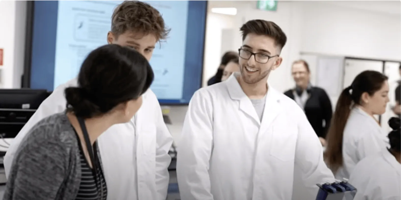 Students in white lab coats speak with an instructor in a lab setting.