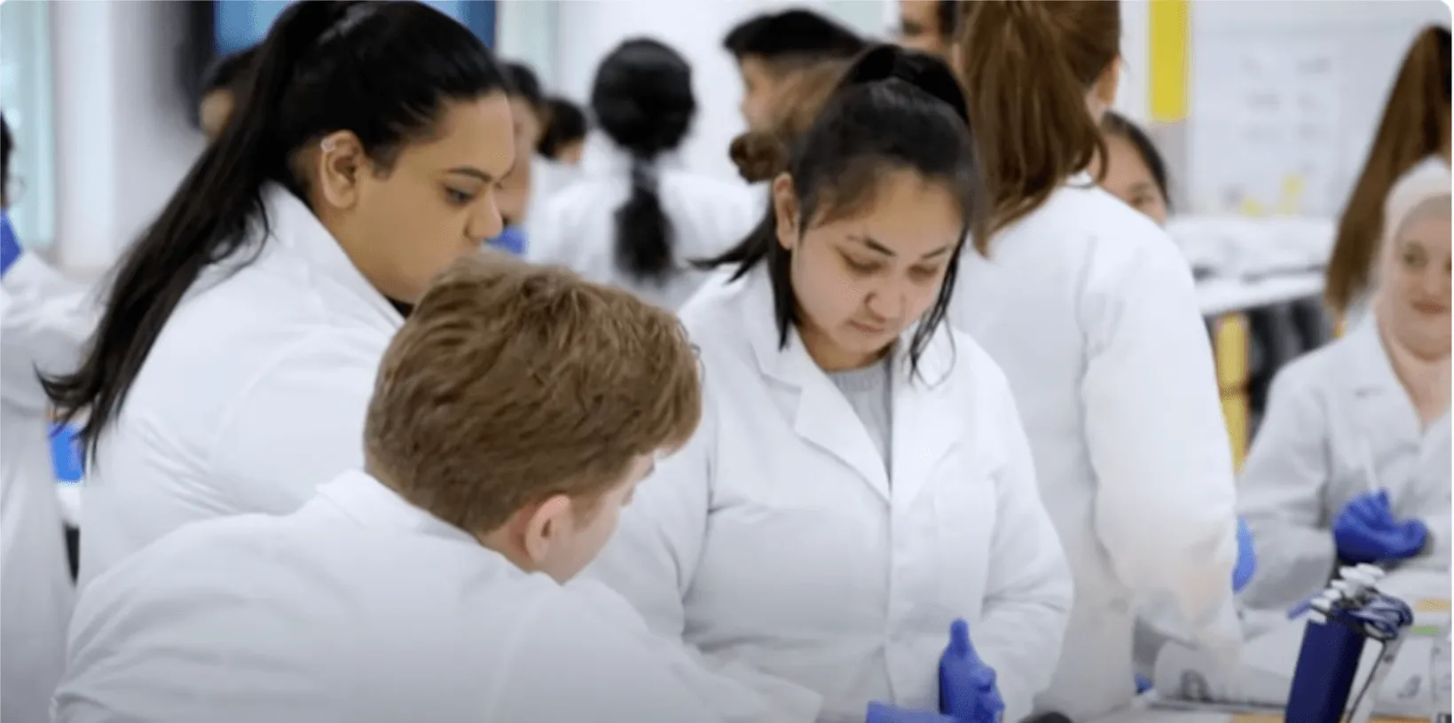 Students in white lab coats work together in a lab environment.