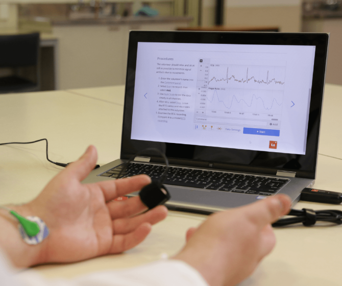 A photograph of a laptop on a table in a physiology lab environment. The software platform Lt is shown on screen, with example physiological data in the data sampling panel. A person's hands are resting in the foreground in front of the laptop, connected to a pulse transducer and a biopotential sensor (via electrodes).