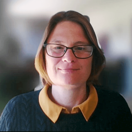 Celia Clarke. She wears glasses and a collared shirt. She has a bob and wears a jumper. She appears against a blurred background, like a Zoom background.