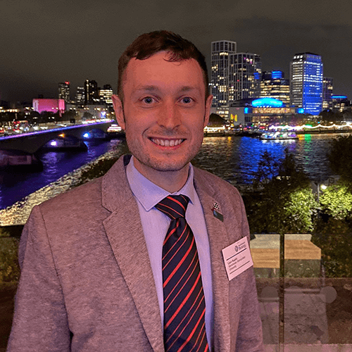 A photograph of a person called Liam Bagley. They have short dark hair and are smiling at the camera. They are wearing a suit and tie, and stand in front of a river and cityscape at night.