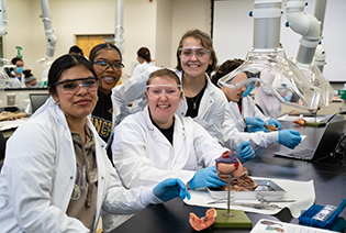 A photograph of four students smiling and wearing lab coats. They are in a lab environment and are participating in a dissection activity.
