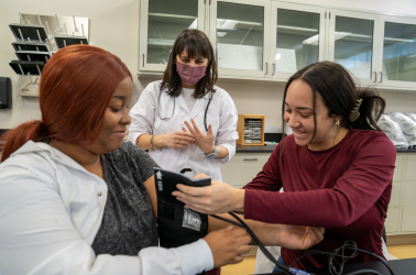 A photo of two students completing a blood pressure exercise in the lab while their educator watches on.