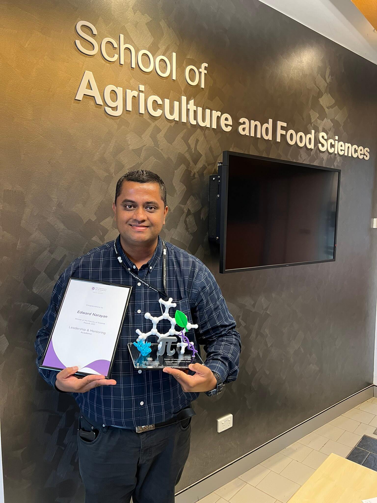 A photo of Dr. Edward Narayan with his UQ Science Award for Leadership & Mentoring . He is standing in front of a sign on the wall saying "School of Agriculture and Food Sciences".