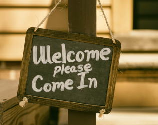 A chalkboard sign hanging on a post. The sign says in white chalk "Welcome please come in".